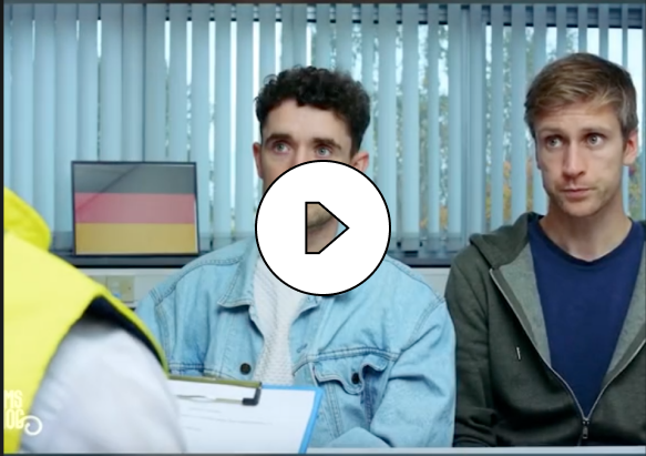 Two men sitting indoors across from a person in a yellow jacket holding a clipboard, with vertical blinds and a German flag in the background.