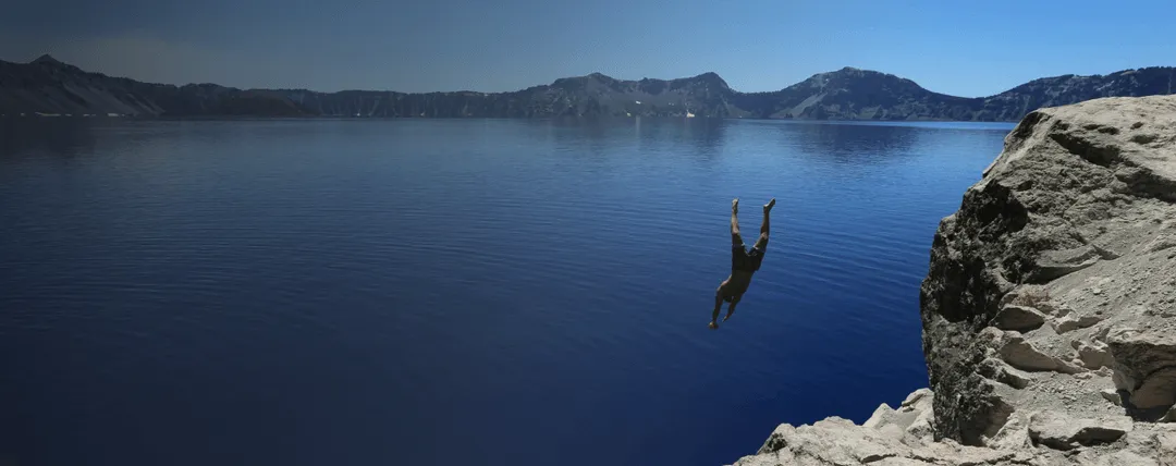 Person diving headfirst into a calm blue lake from a rocky cliff with mountains in the background.