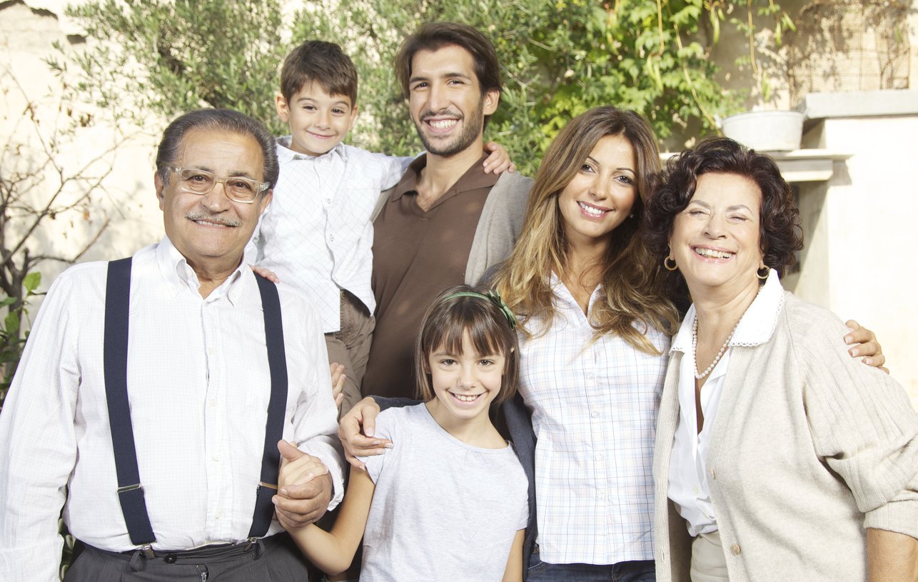 Smiling multi-generational family of six standing outdoors with greenery in background.