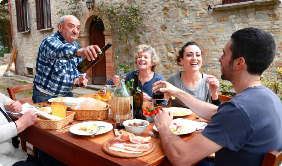 Four adults enjoying an outdoor meal at a wooden table with wine and Italian-style food, seated near a stone building.