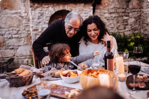 Older man, woman, and child smiling and interacting at an outdoor dining table with food and wine.