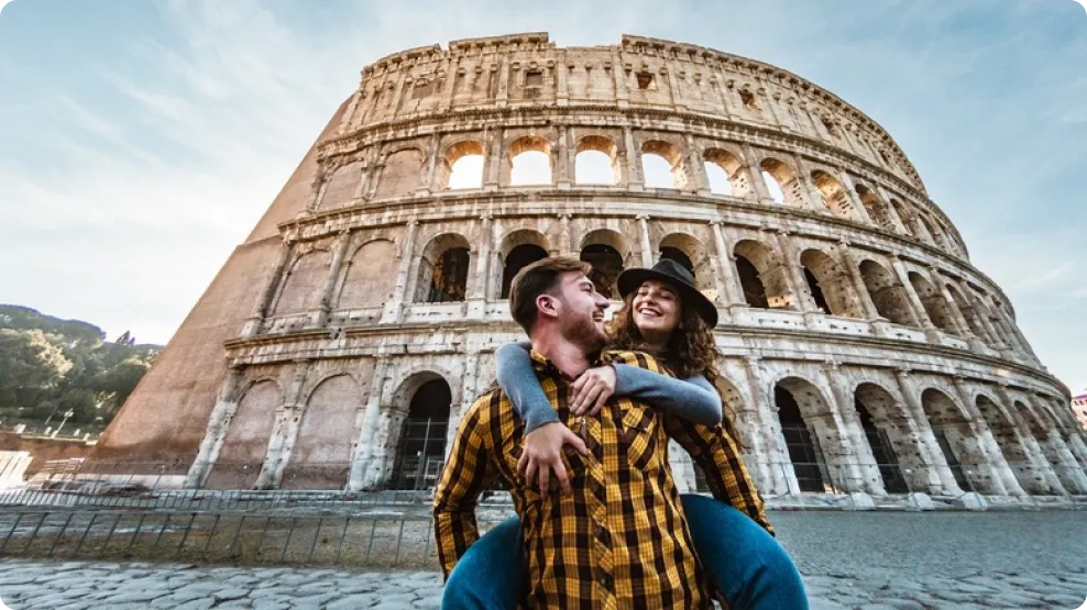 Smiling young couple with the man giving the woman a piggyback in front of the ancient Roman Colosseum.