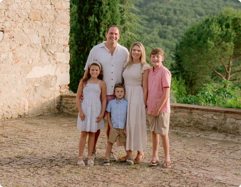 Smiling family of five posing outdoors on a stone path with greenery and trees in the background.