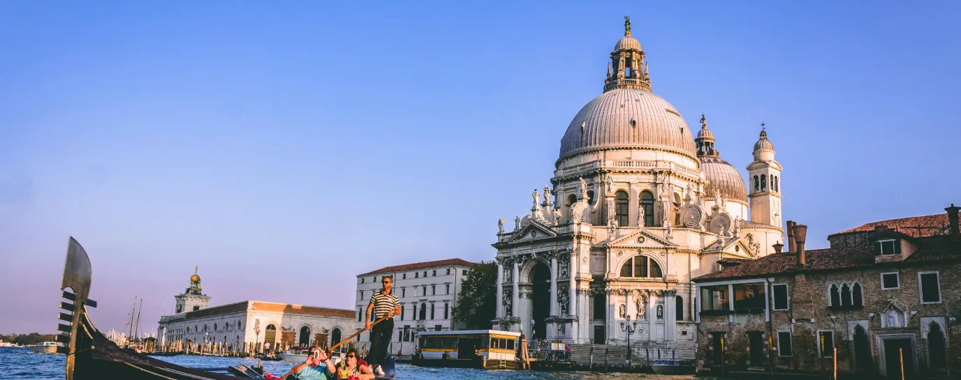 Gondola with passengers and a gondolier on a canal in Venice in front of the Santa Maria della Salute basilica.
