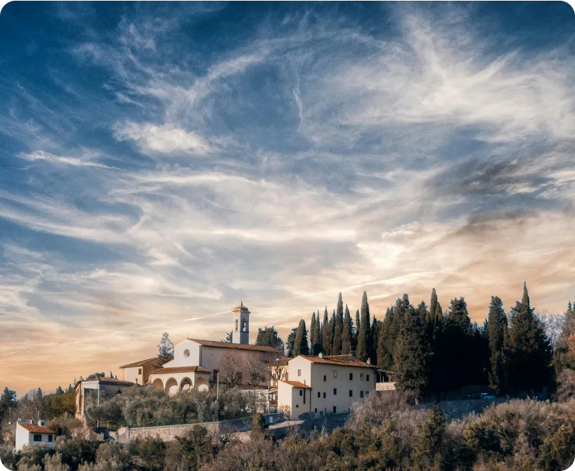 Hilltop estate with Mediterranean-style buildings and tall cypress trees under a partly cloudy sky at sunset.