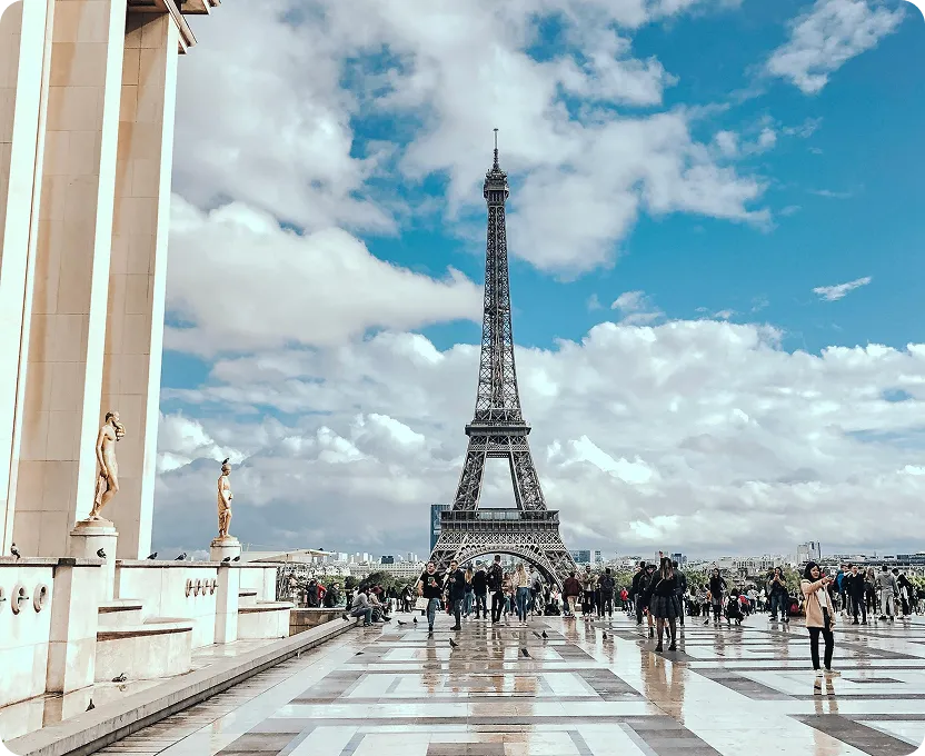 Crowd of people walking and taking photos on a tiled plaza with the Eiffel Tower and partly cloudy blue sky in the background.