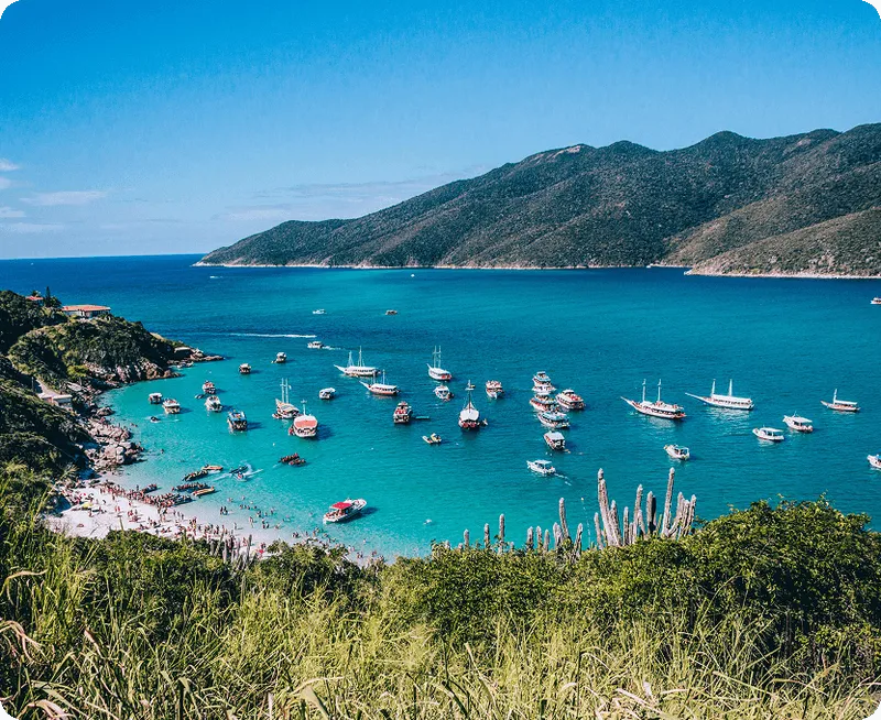 Scenic bay with turquoise water, numerous boats anchored near a sandy beach, lush green hills, and clear blue sky.