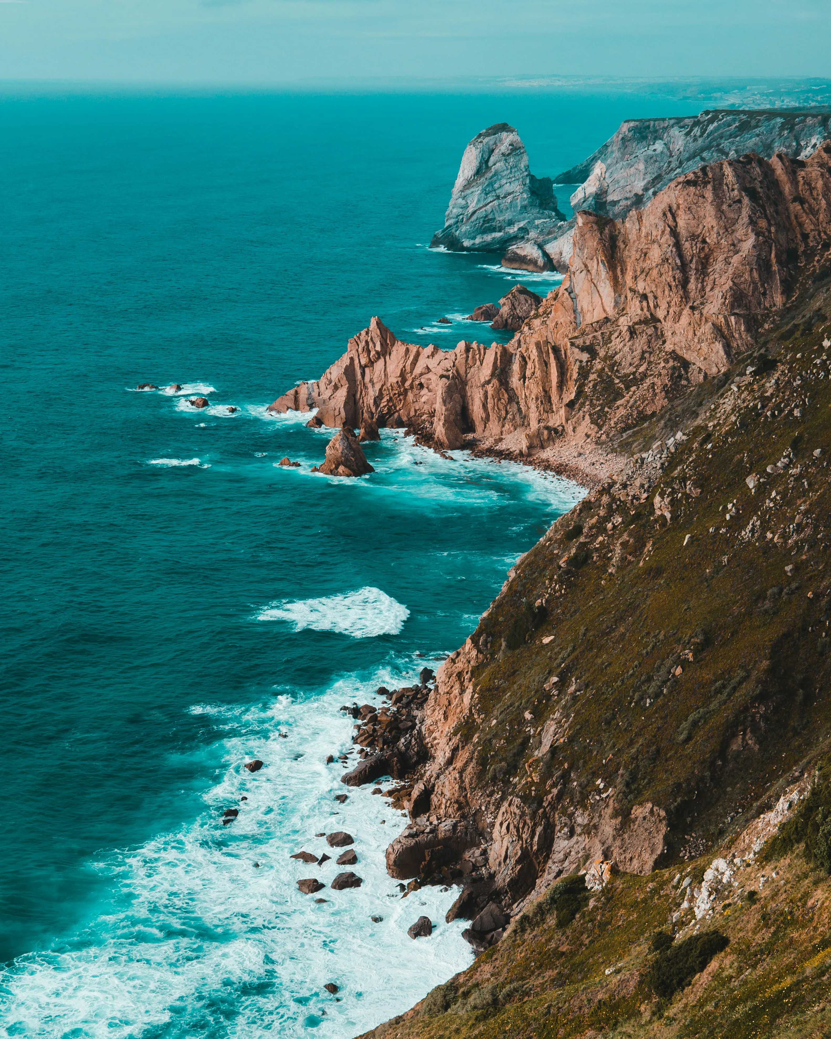 Rocky coastline with steep cliffs and waves crashing against the shore under a clear sky.