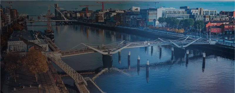 Modern pedestrian bridge with unique angular supports spanning a river in a cityscape at dusk with buildings and cranes in the background.
