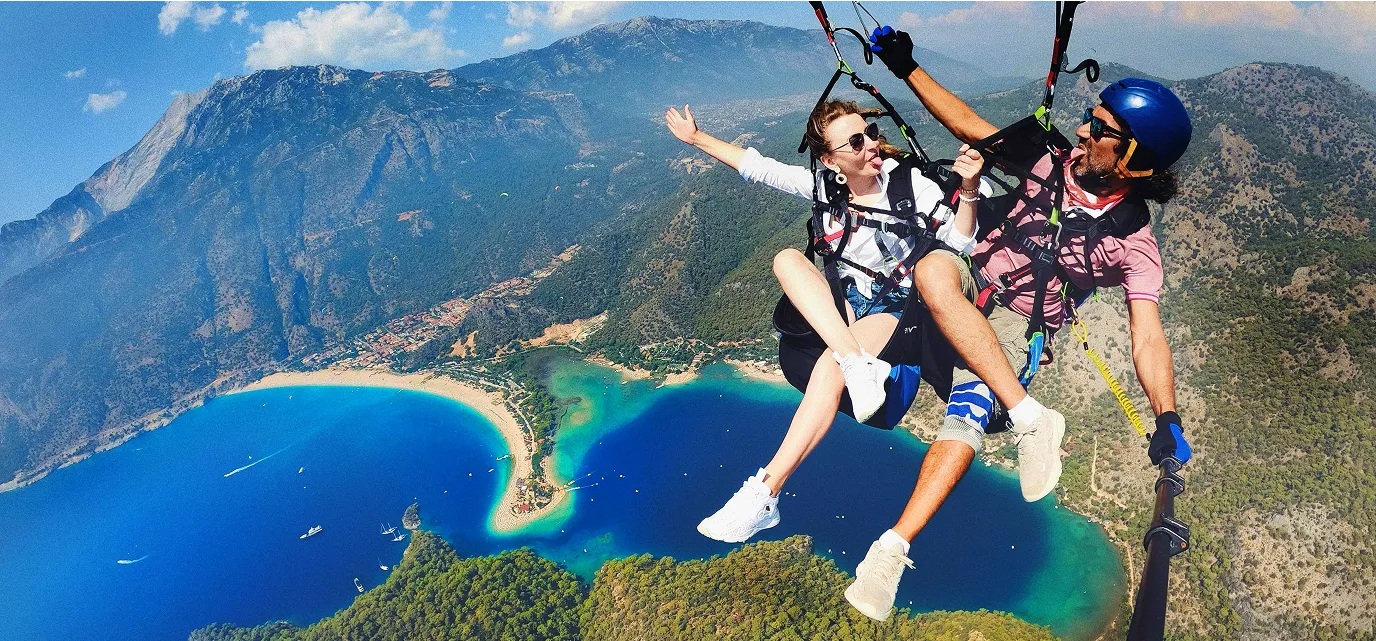 A man and woman tandem paragliding over a turquoise bay with mountainous landscape in the background.