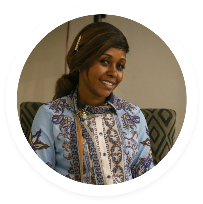 Smiling woman with a hairpin in ponytail wearing a patterned light blue and white top, seated on a patterned chair.