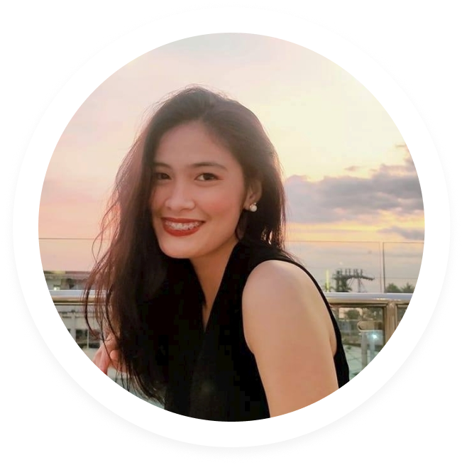 Smiling woman with long dark hair and pearl earrings wearing a black sleeveless top, with a sunset sky in the background.