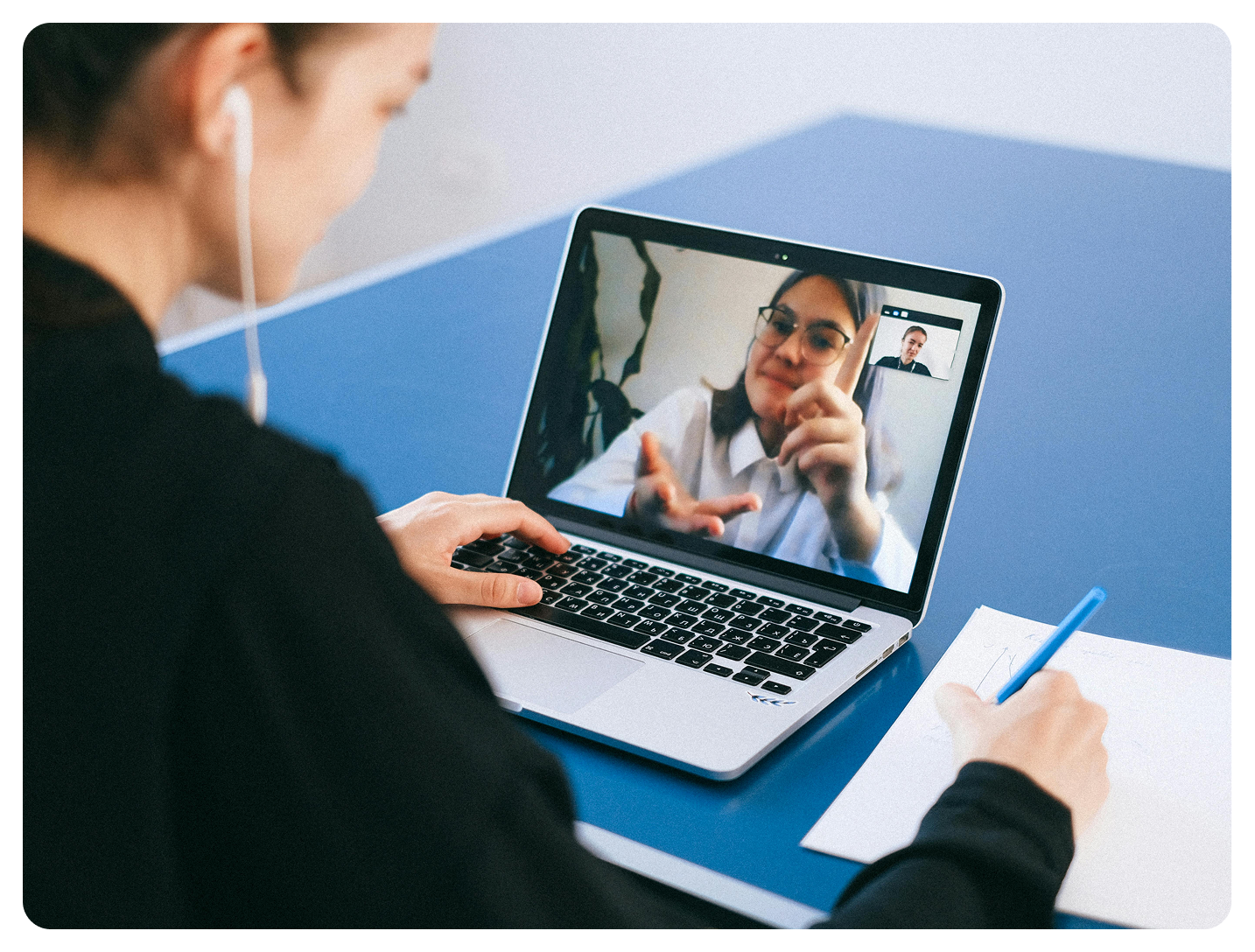 Person wearing earphones engages in video call on laptop while taking notes on paper with a blue pen.