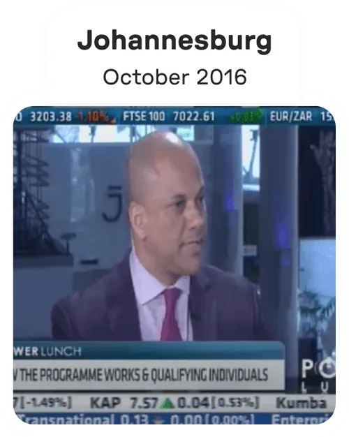 Man in a suit and red tie speaking during a televised interview with financial stock tickers visible at the top and bottom of the screen.