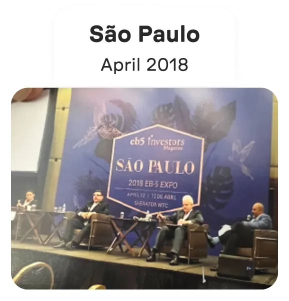 Panel discussion with four men seated on stage at the São Paulo 2019 EB-5 Expo in front of a blue backdrop displaying event details.