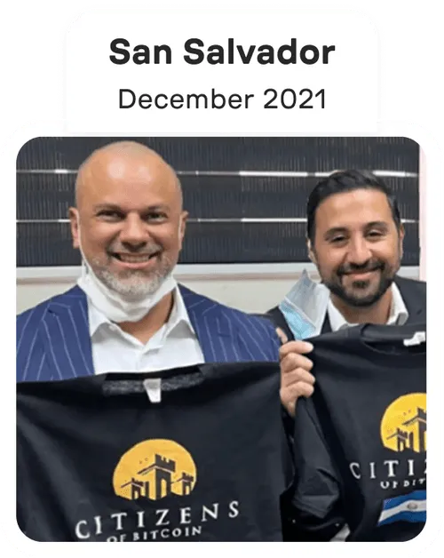 Two men smiling and holding black t-shirts with the 'Citizens of Bitcoin' logo in front of a dark background.