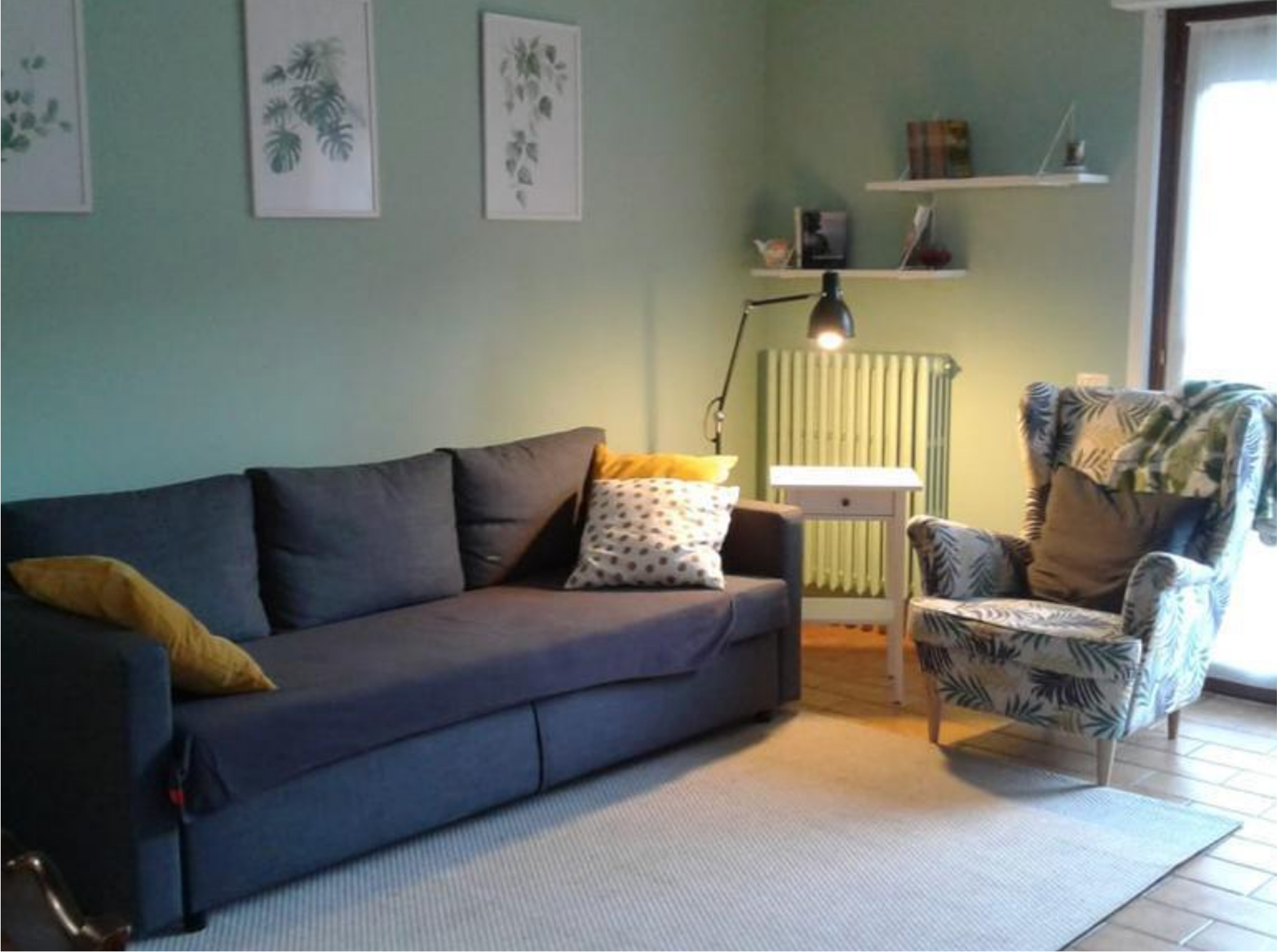 Living room with a dark gray sofa with cushions, patterned armchair, white side table with a black lamp, and botanical wall art.