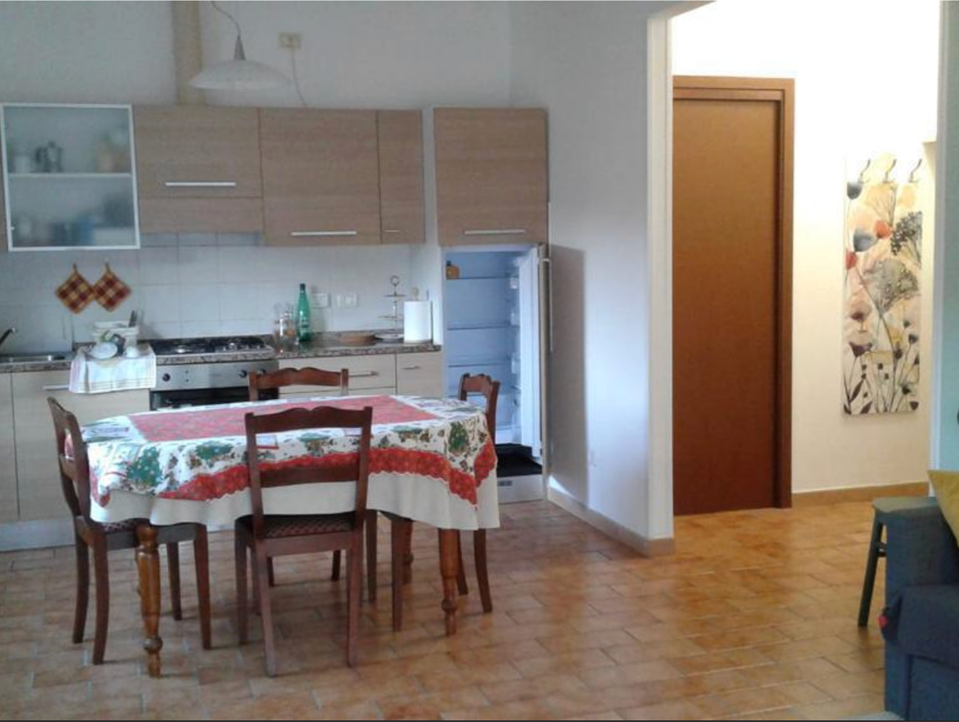 Kitchen and dining area with wooden table covered by a floral tablecloth, four wooden chairs, light wood cabinets, tiled floor, and an open refrigerator.