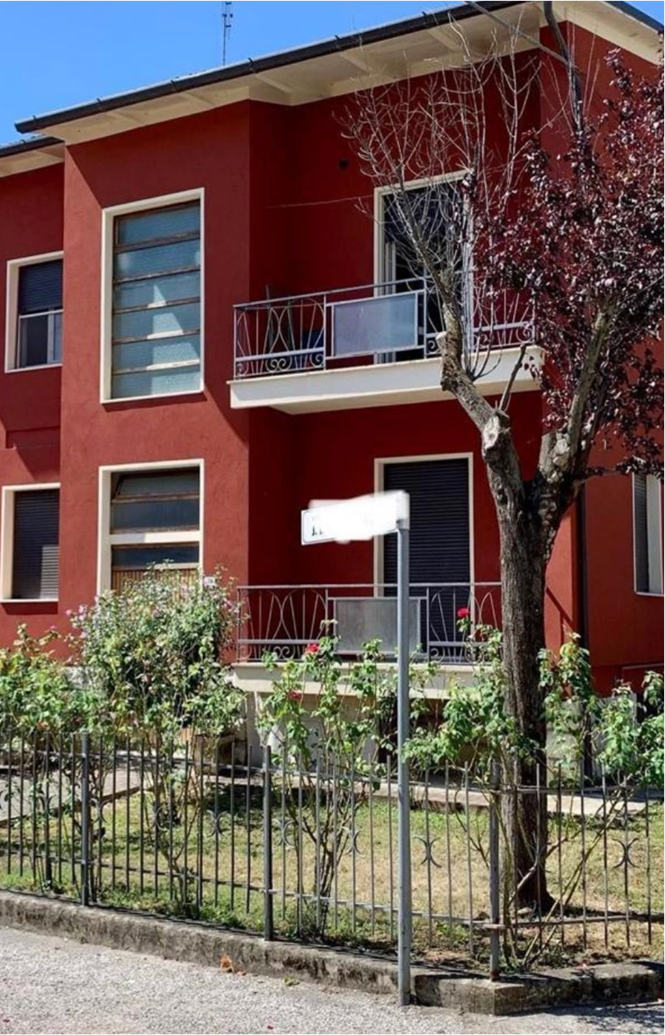 Two-story red house with white trim, balconies with metal railings, a fenced garden, and a tree in front under clear blue sky.
