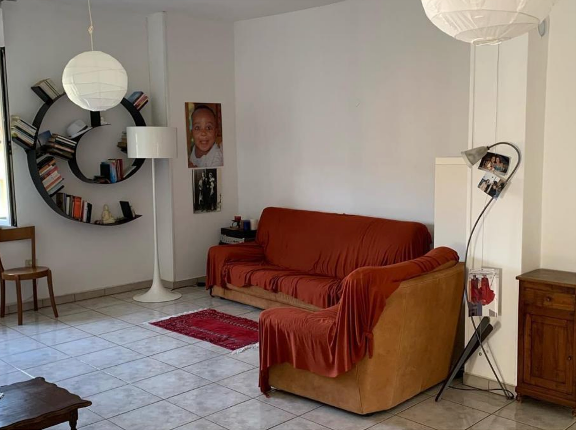 Living room with tan sofa and armchair covered in rust-colored throws, circular black wall-mounted bookshelf, floor lamp, wooden chair, photos on walls, and tiled floor.