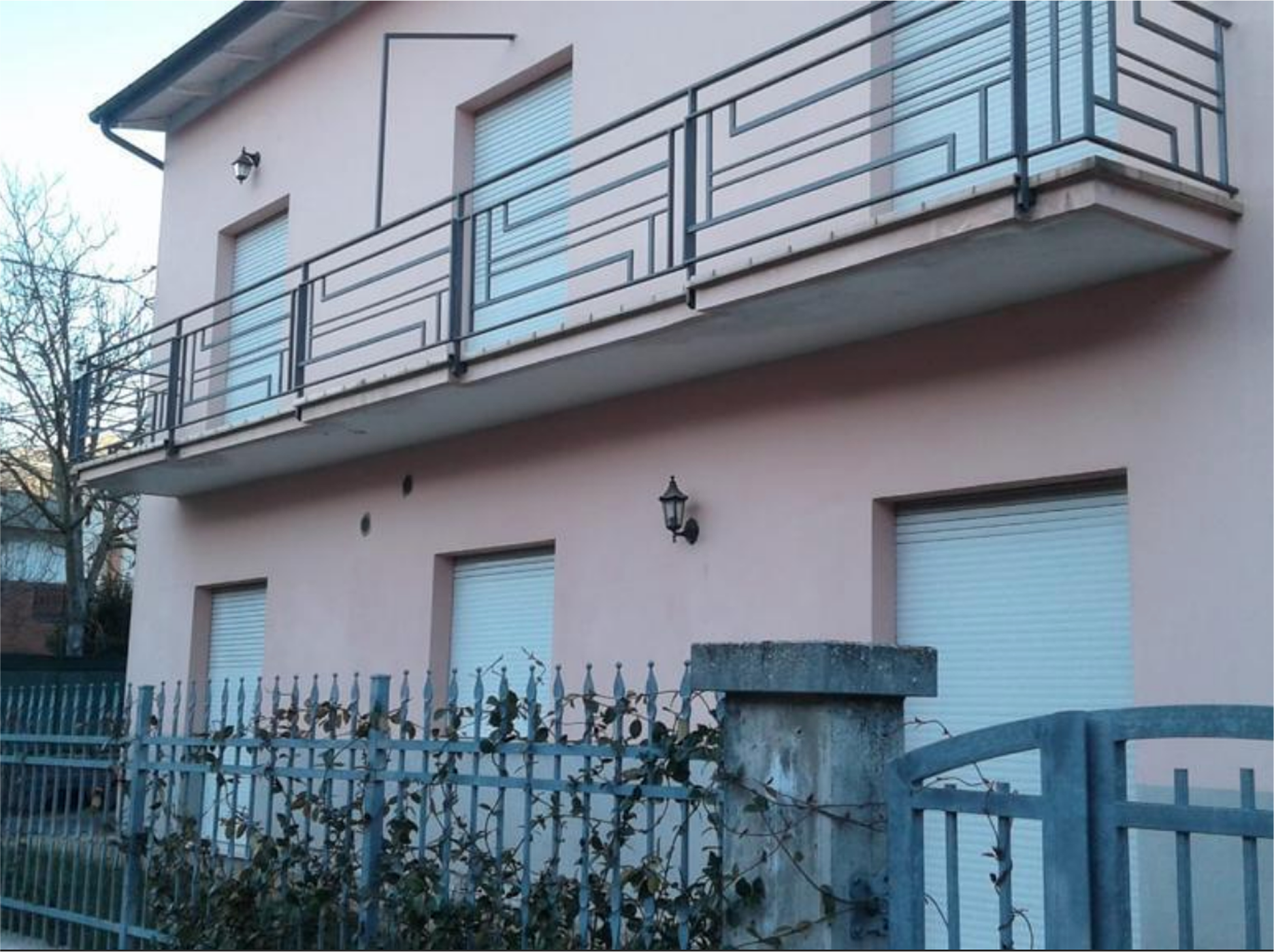 Two-story pink house with closed white window shutters, a metal balcony railing, and a metal gate with climbing vines.