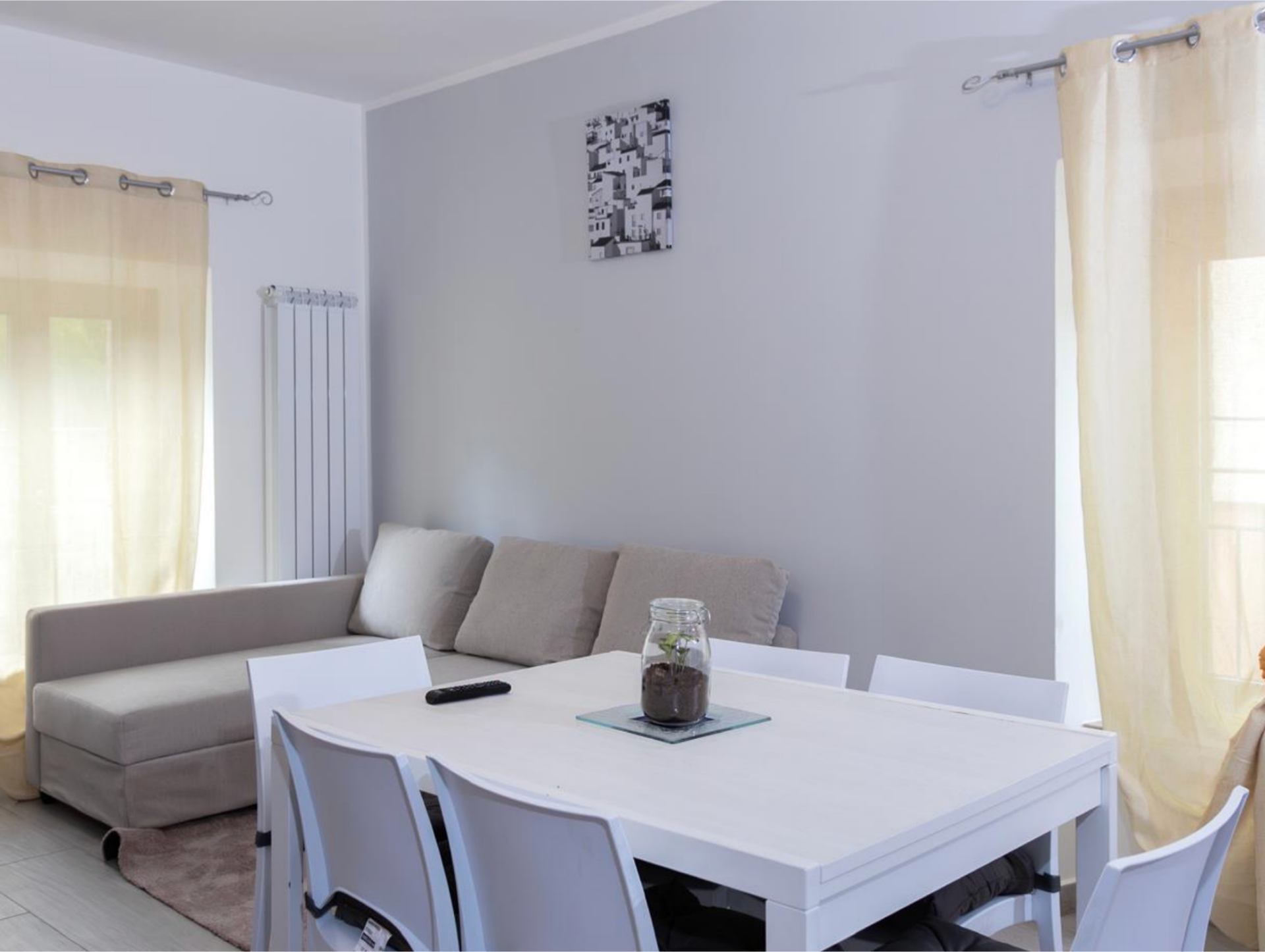 Bright dining area with a white table, six white chairs, a small glass jar with a plant centerpiece, and a beige sofa with cushions against the wall.