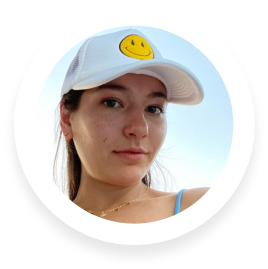 Young woman wearing a white cap with a yellow smiley face, outdoors with clear sky background.