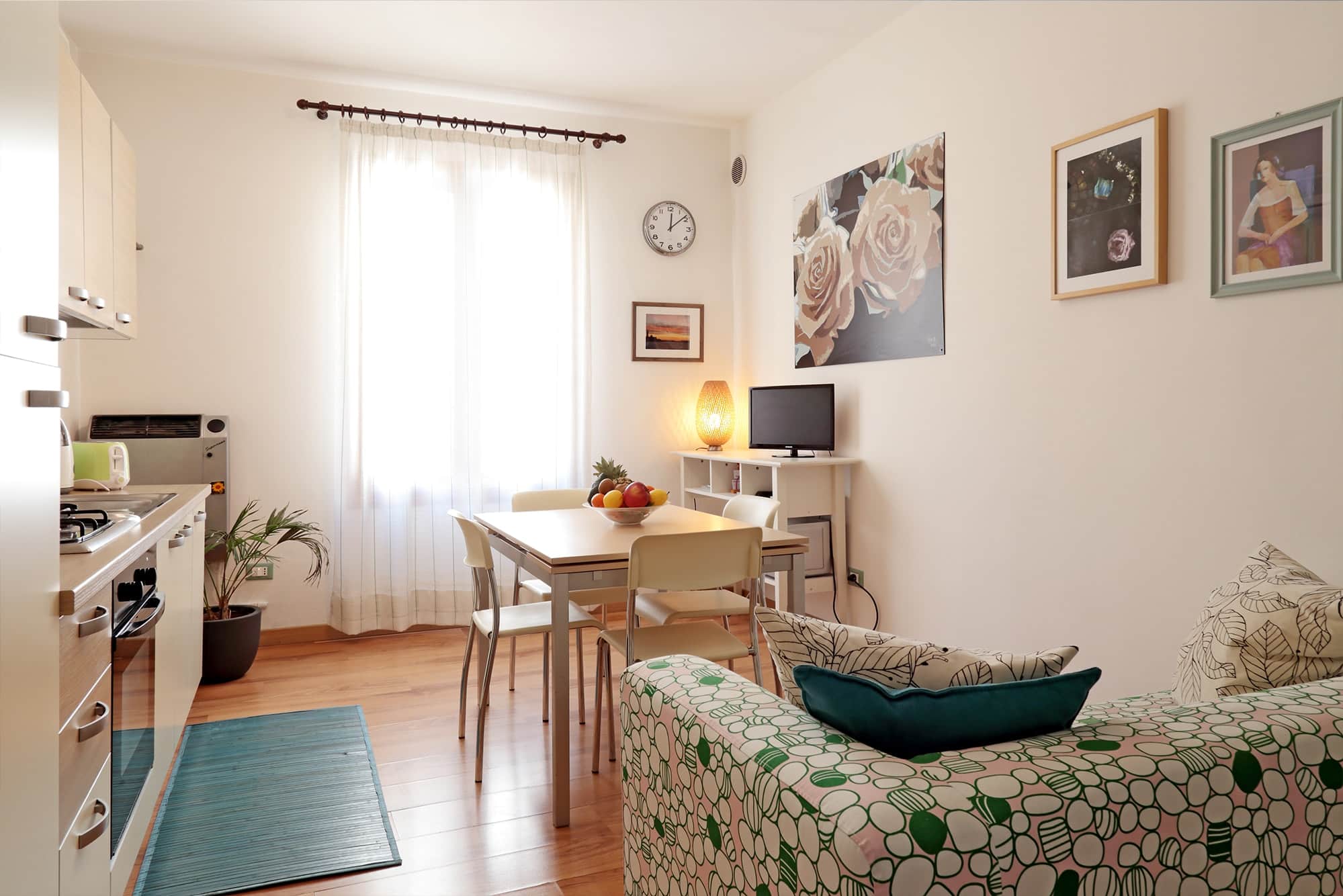 Bright kitchen and dining area with wooden floor, table with four chairs and fruit bowl, patterned sofa, and wall art.