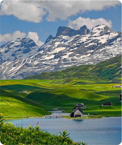 Snow-capped mountains behind rolling green hills and a lake with small wooden houses on its shore.