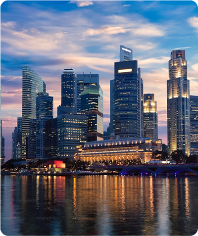 City skyline at dusk with illuminated skyscrapers reflecting on calm water under a partly cloudy sky.