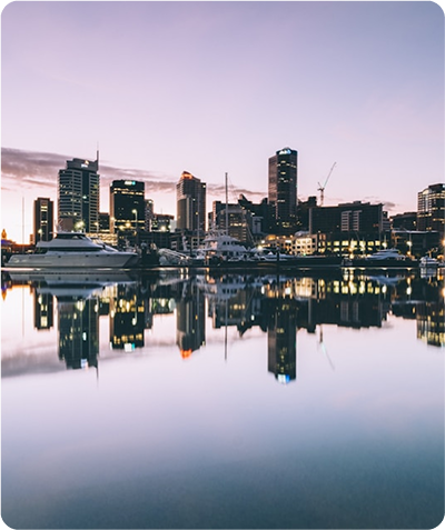 City skyline at dusk reflected on calm water with boats docked in the harbor.