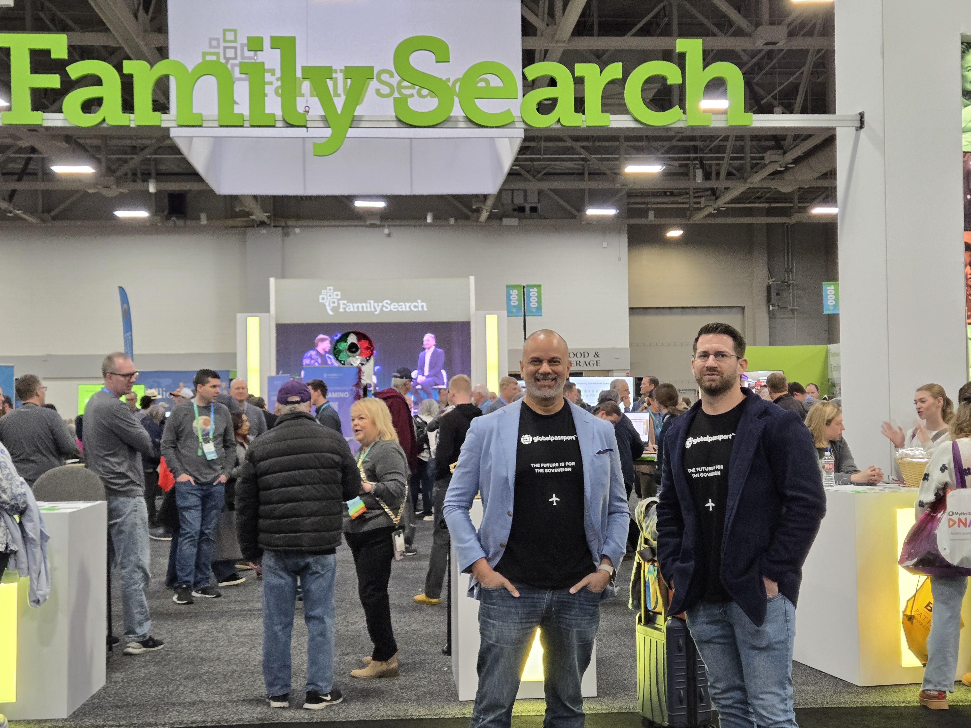 Two men wearing 'globalpassport' shirts stand in front of a FamilySearch booth at a crowded convention.