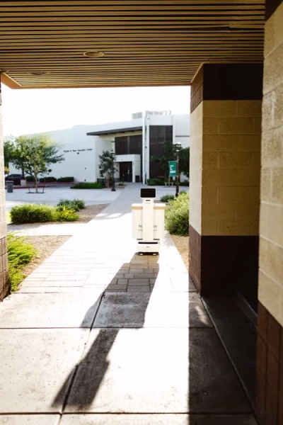 A white delivery robot with a black screen on top traveling on a sidewalk toward a modern building entrance.