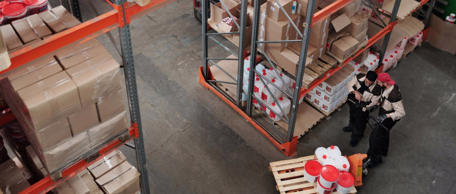 Two warehouse workers checking inventory while standing next to stacked pallets of boxes and containers in a storage facility.