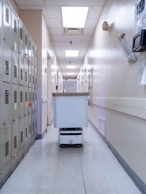 Autonomous delivery robot moving down a hallway with lockers on the left and papers on the wall to the right.
