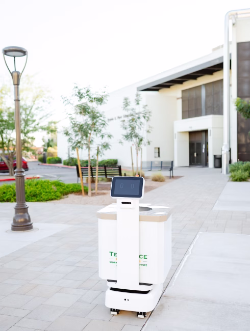 Autonomous delivery robot with a digital display navigating an outdoor walkway near a building and lamp post.