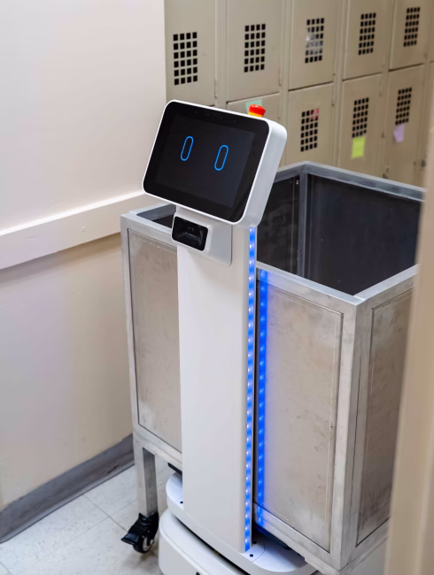 White service robot with digital face and blue LED lights beside a metal waste bin in an indoor setting with lockers in the background.