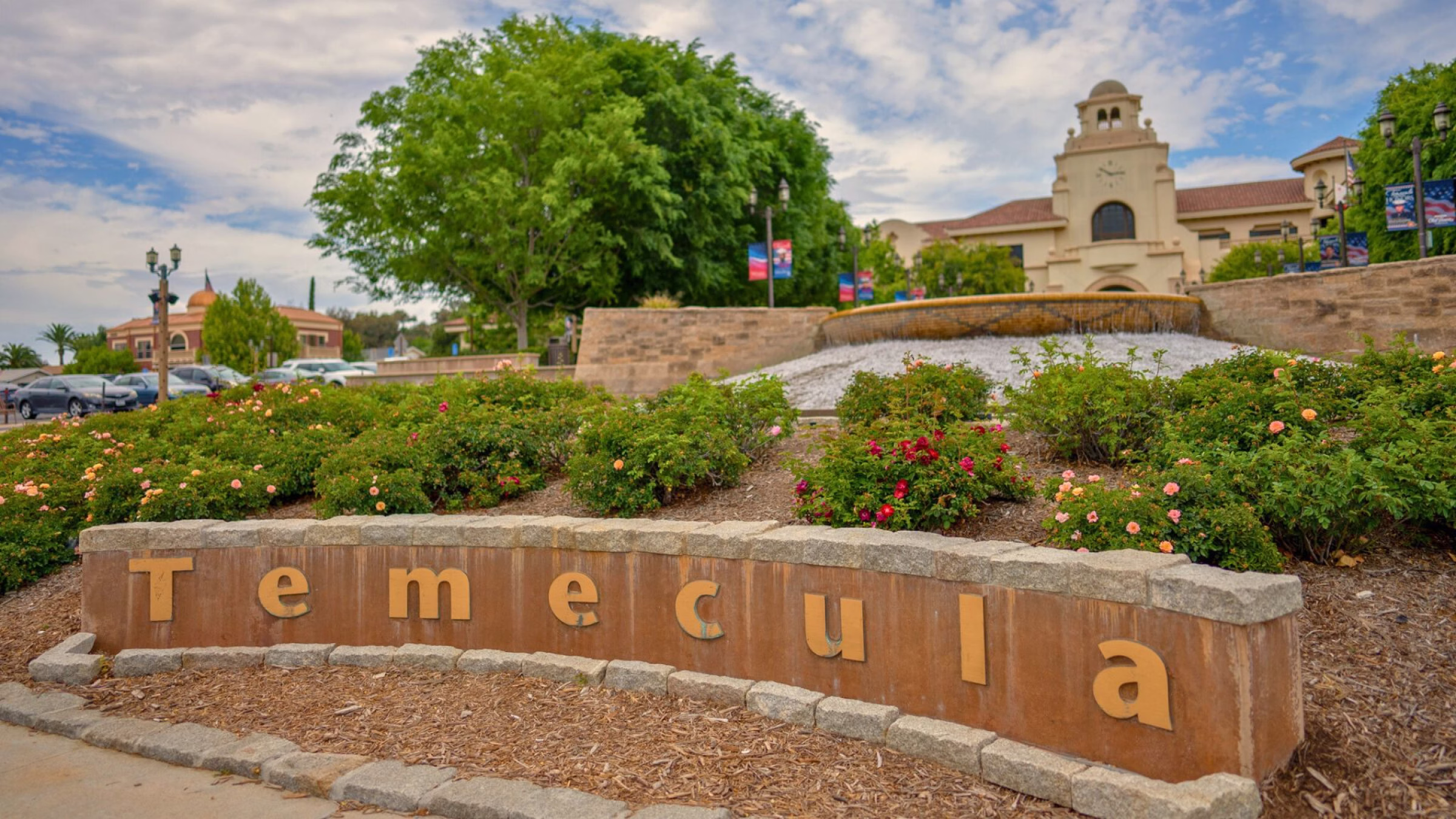 Stone sign with large letters spelling 'Temecula' in front of flower bushes, a cascading fountain, and a clock tower building.