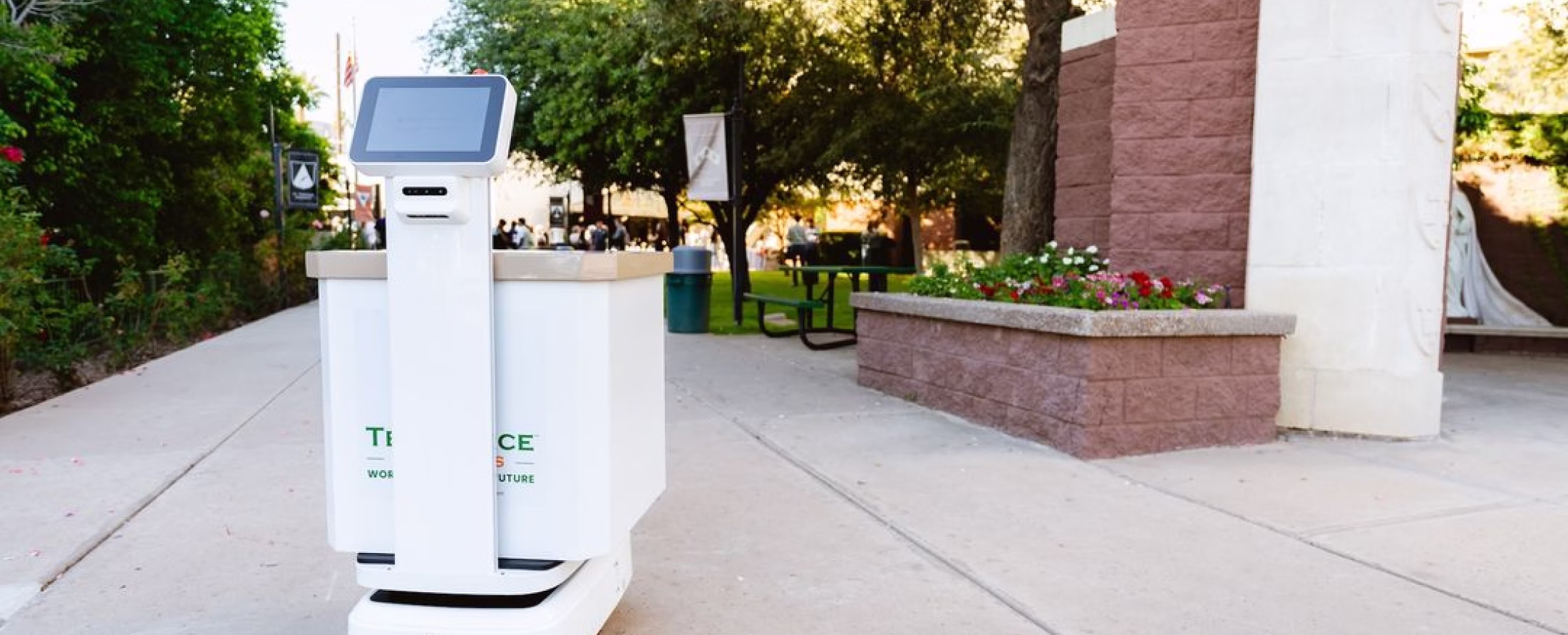 White autonomous delivery robot with a screen on top moving on a sidewalk near a campus area with trees and benches.