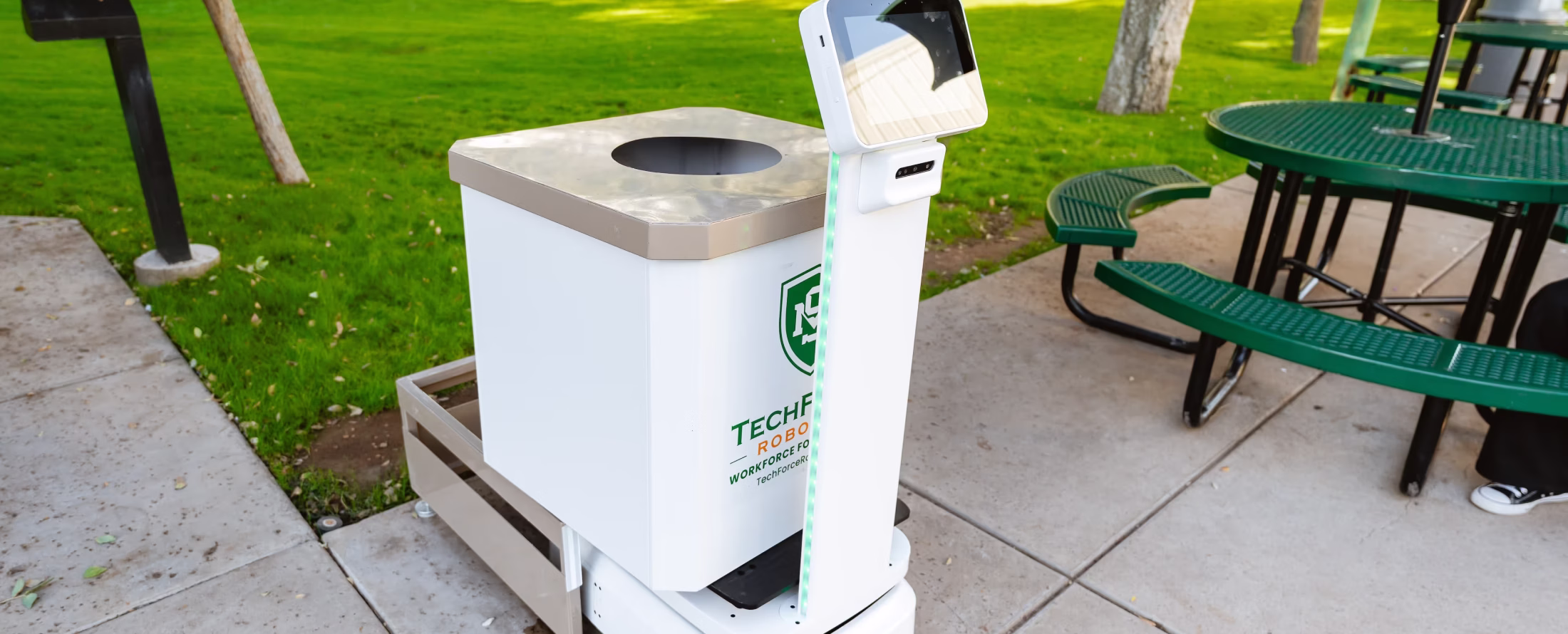 Small white robotic waste bin with a touch screen on a sidewalk next to green picnic tables and grass.
