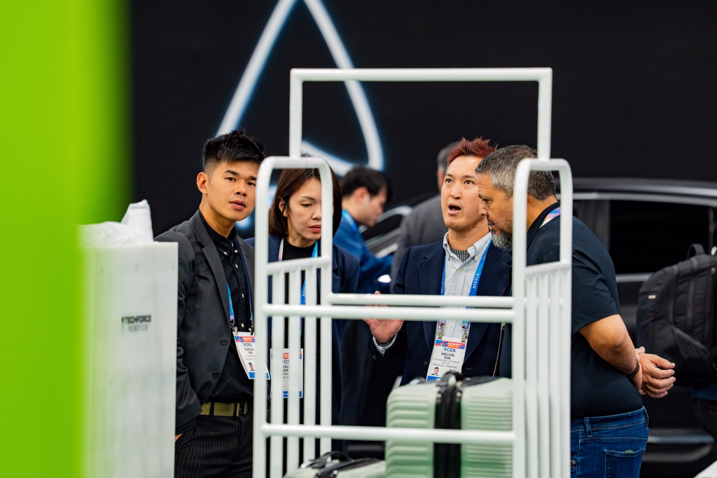 Four people engaged in conversation at an indoor event, seen through a white luggage cart with suitcases.