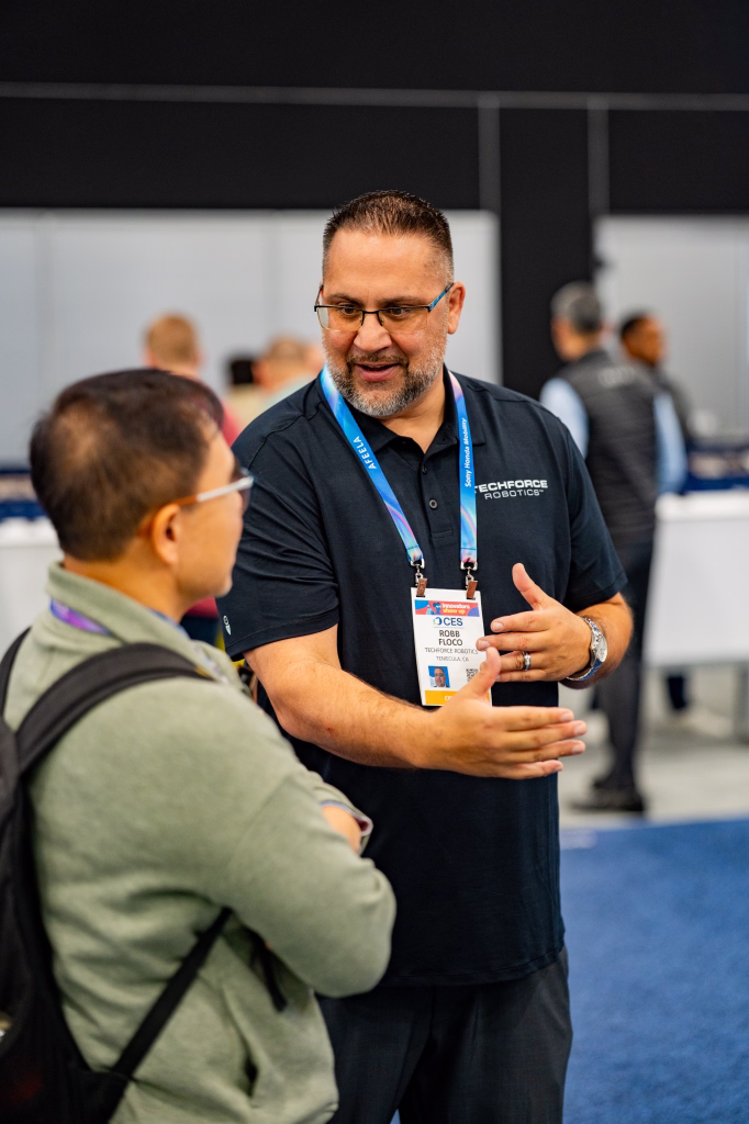 Man wearing a Techforce Robotics shirt and conference badge engaging in conversation with another man in an indoor event setting.