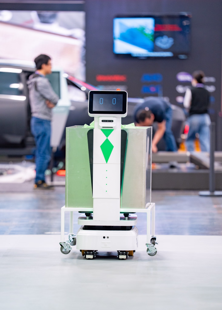 White delivery robot with digital face and green tie design on a warehouse floor with people blurred in the background.