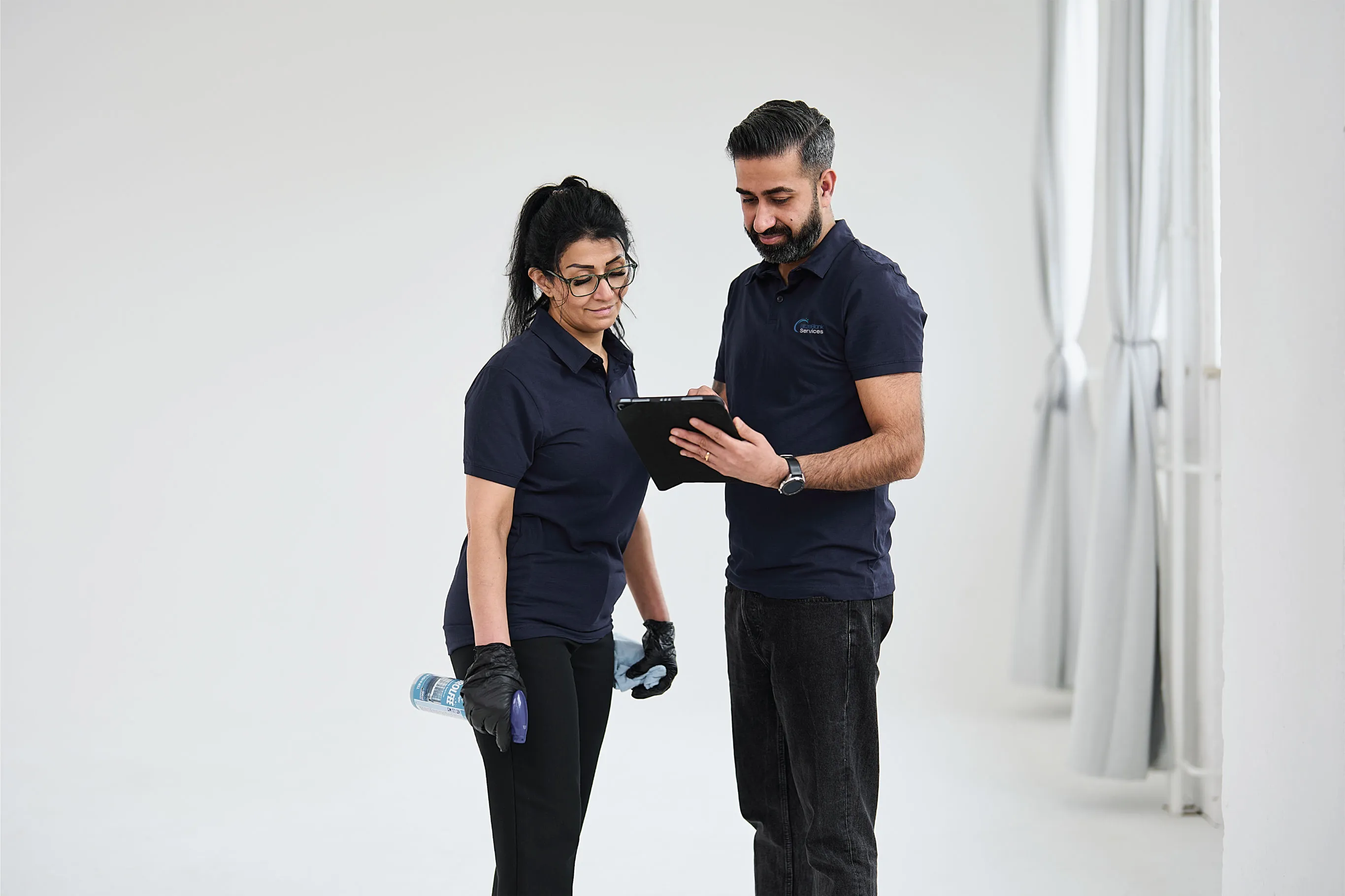 Zwei Personen in dunkelblauen Poloshirts stehen vor weißem Hintergrund, eine hält ein Tablet, die andere eine Flasche Reinigungsmittel und trägt Handschuhe.