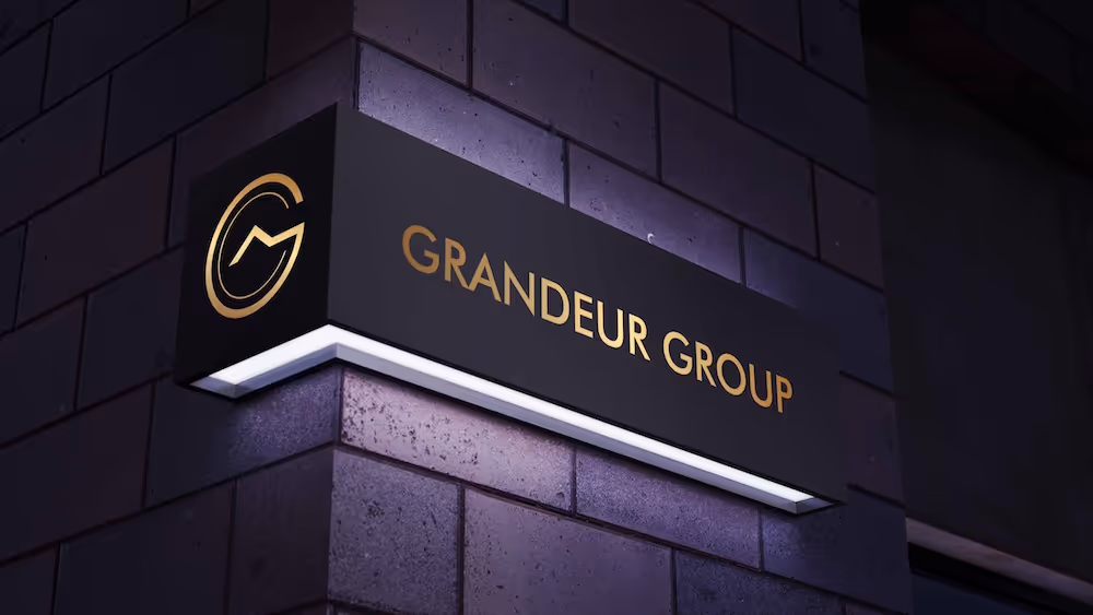 Illuminated black sign mounted on a brick wall corner displaying the text 'GRANDEUR GROUP' and a gold circular logo.