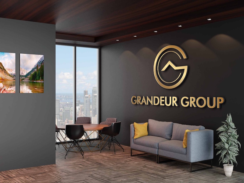 Modern office lounge area with grey sofa, yellow cushions, wooden ceiling, and Grandeur Group gold logo on dark wall next to large windows overlooking a cityscape.
