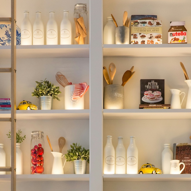 Kitchen shelves with white bottles, wooden cooking utensils in jars, plants, a glass jar with red ornaments, cookbooks, and small yellow toy cars.