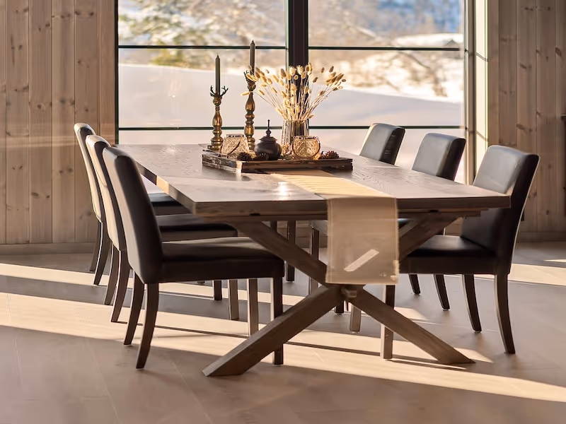 Wooden dining table with six black chairs in a sunlit room, decorated with a beige table runner and a centerpiece of candlesticks and dried flowers.