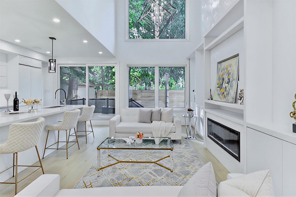 Bright modern living room with white furniture, glass coffee table, kitchen island with bar stools, large windows, and a fireplace with artwork above it.