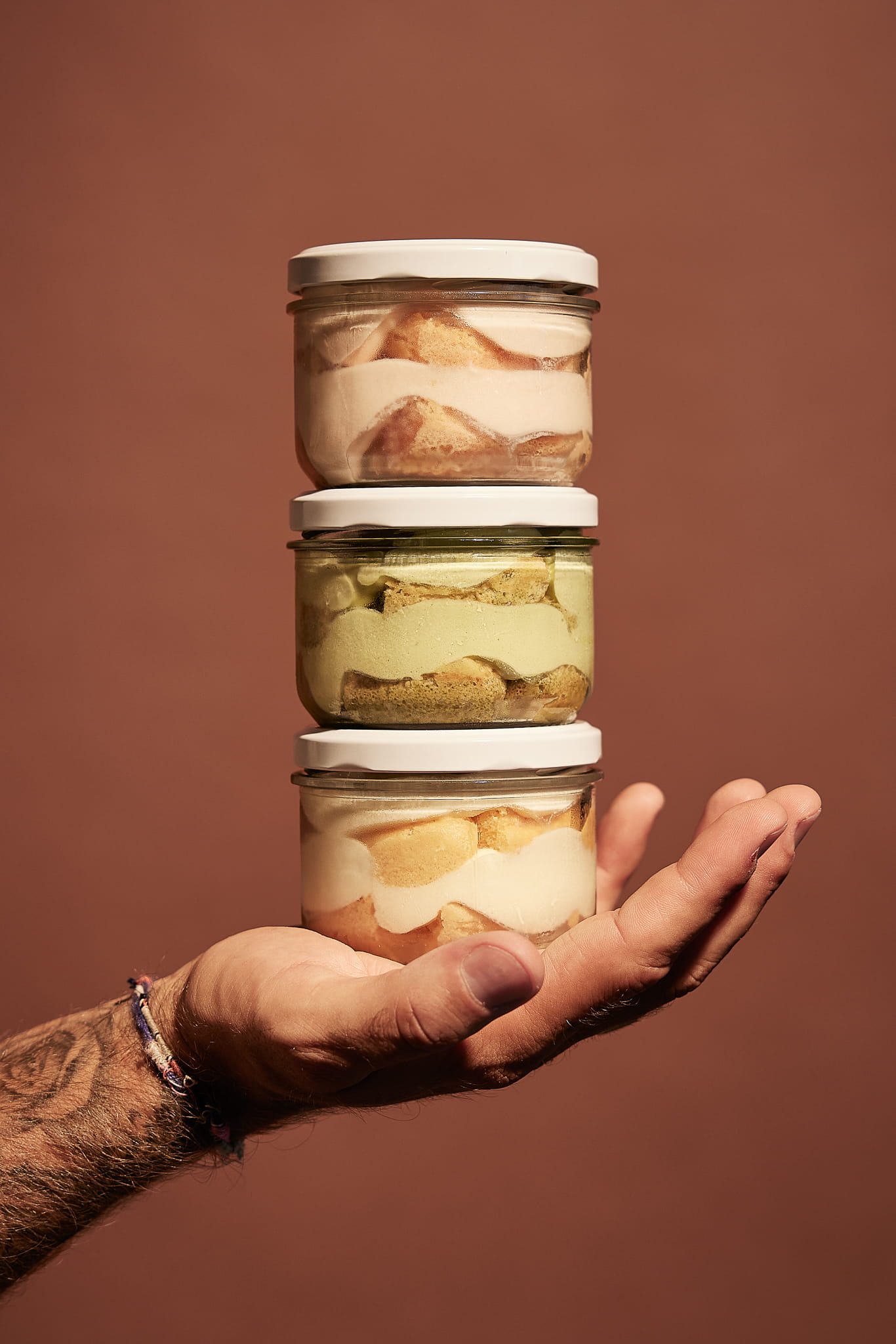 Hand holding a stack of three small glass jars filled with layered tiramisu desserts in different flavors.